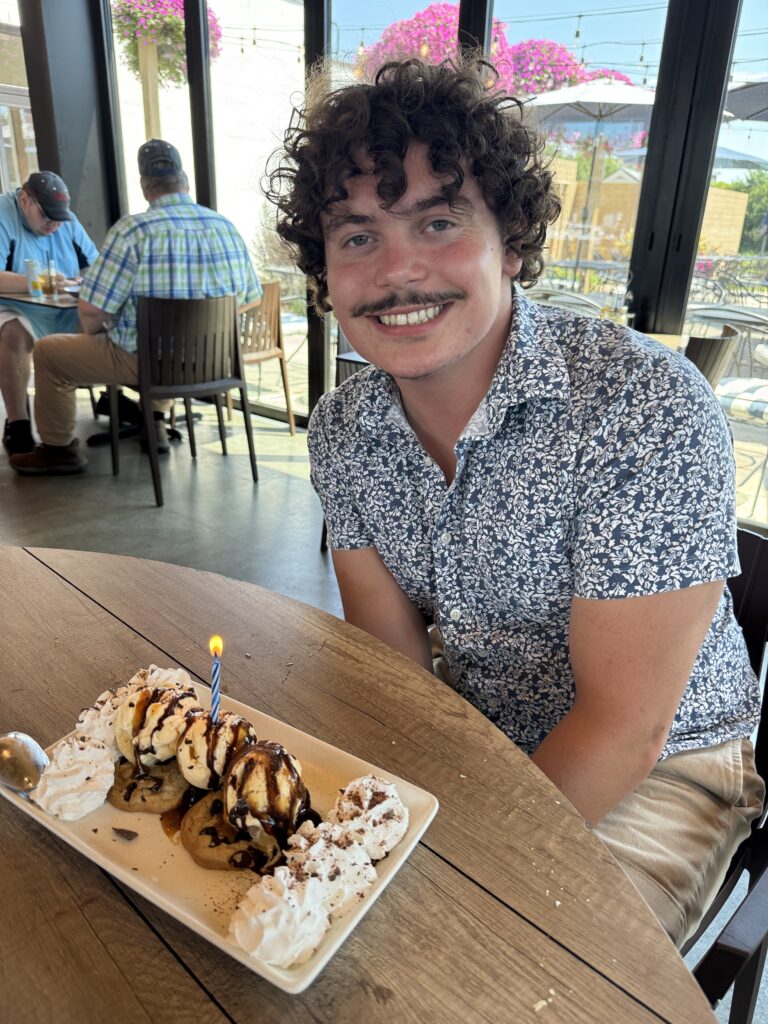 Photo of Aiden Johnson in a blue and white shirt with a smiling expression at a restaurant. There are windows sprawling on the walls with sunny weather outside. On the table is an ice cream treat, and a lit candle.
